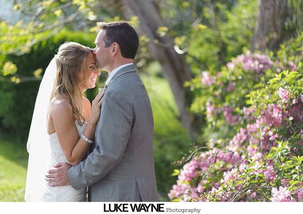 Bride and groom embracing, man kisses woman's forehead outdoors near flowering bushes.
