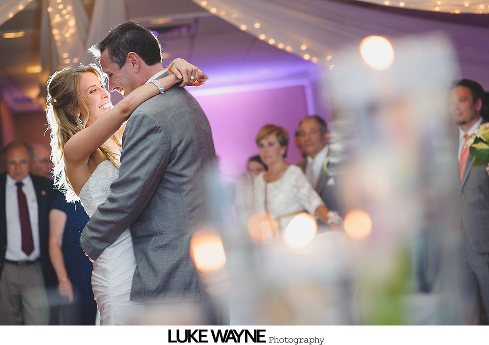 Bride and groom dance at reception. They embrace, smiling, under string lights. Guests watch in background.
