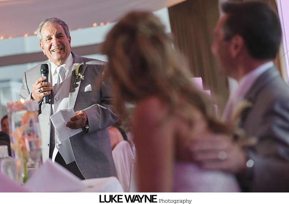 Man giving speech at wedding reception, holding microphone and papers. Bride and groom in soft focus.