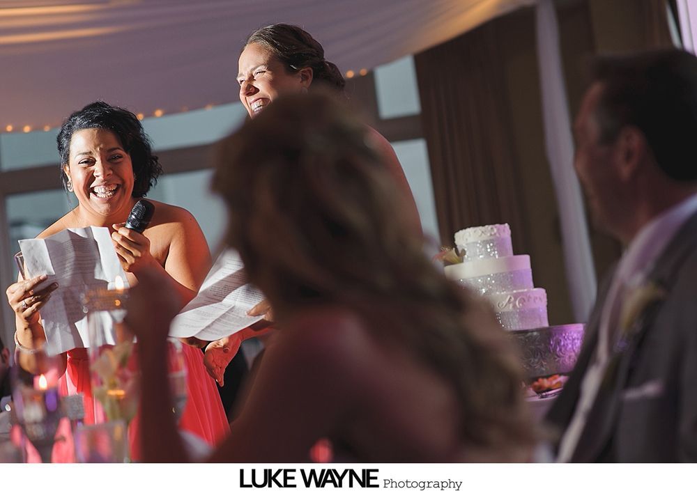 Woman giving a wedding speech, laughing, holding papers, with a cake in the background.