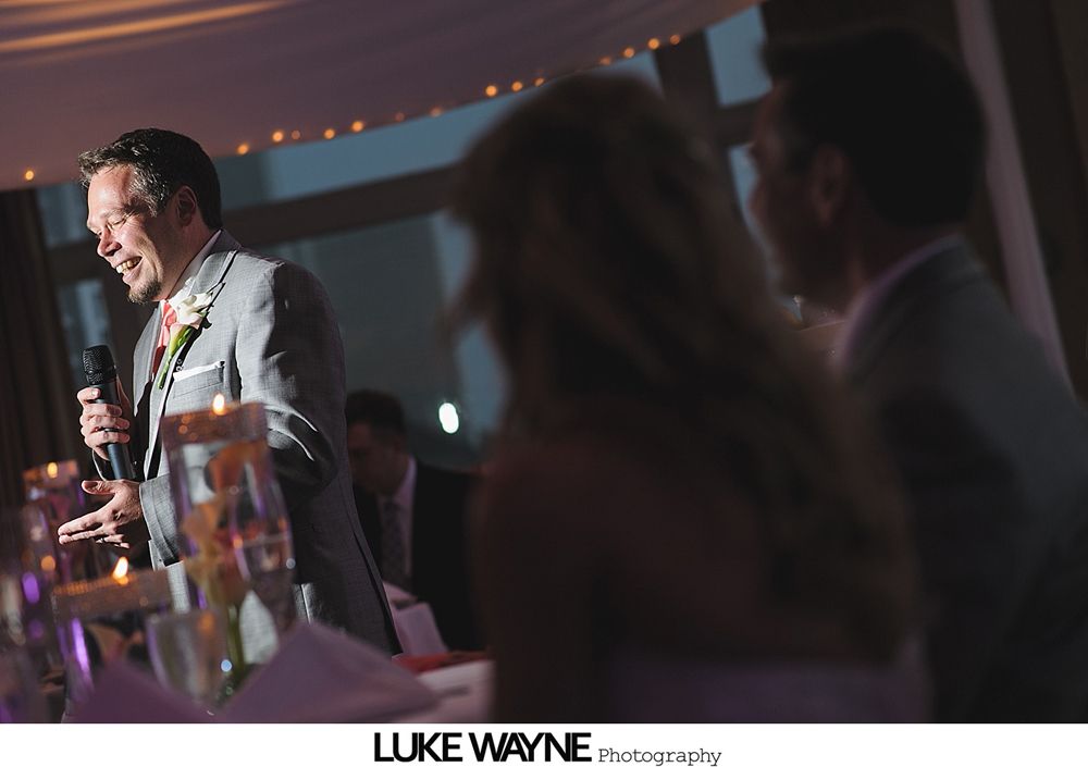 Man in gray suit giving a speech at a wedding, smiling, holding a microphone.