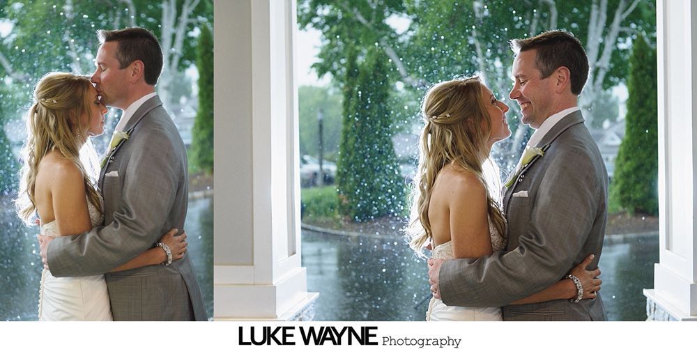 Wedding couple embracing by a window, with rain outside. Man kisses woman's forehead, both smiling.
