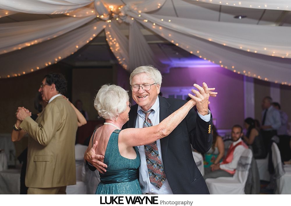 Couple dances at a wedding reception; string lights and white fabric decorate the ceiling.