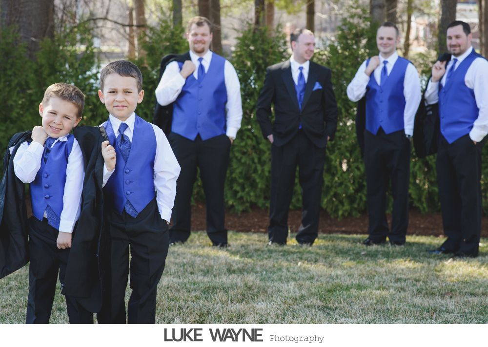 Men in formal attire, including royal blue vests and black suits, pose outdoors. Two young boys in similar attire stand in front.