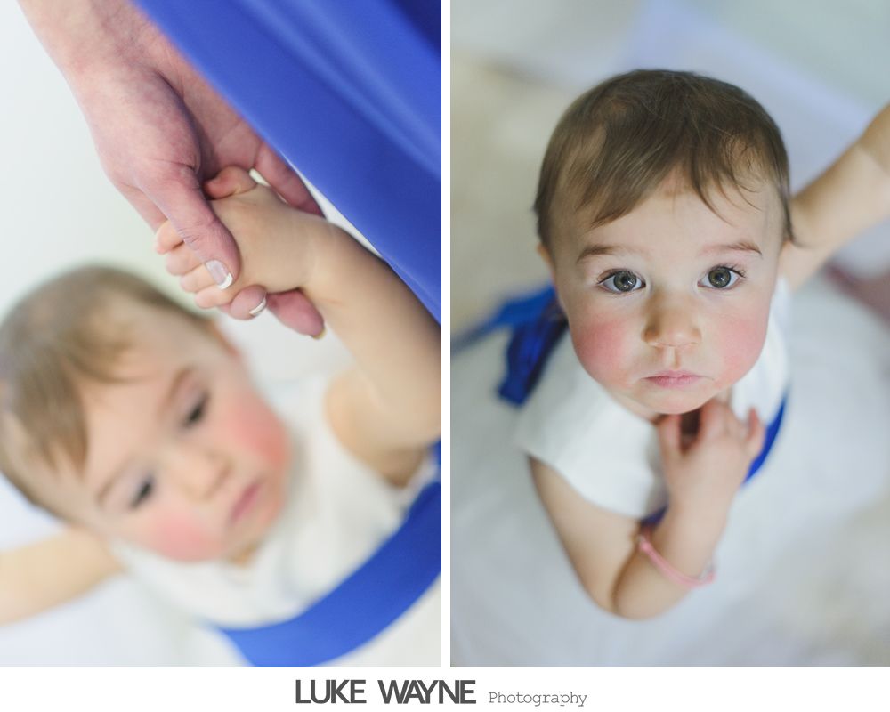 A baby in white dress with blue sash is held by a hand. Looking up with rosy cheeks.