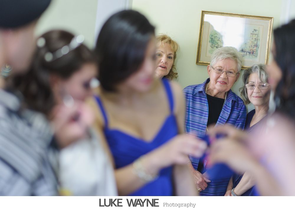 People gathered in a room, watching a woman in a blue dress. A painting hangs on the wall.