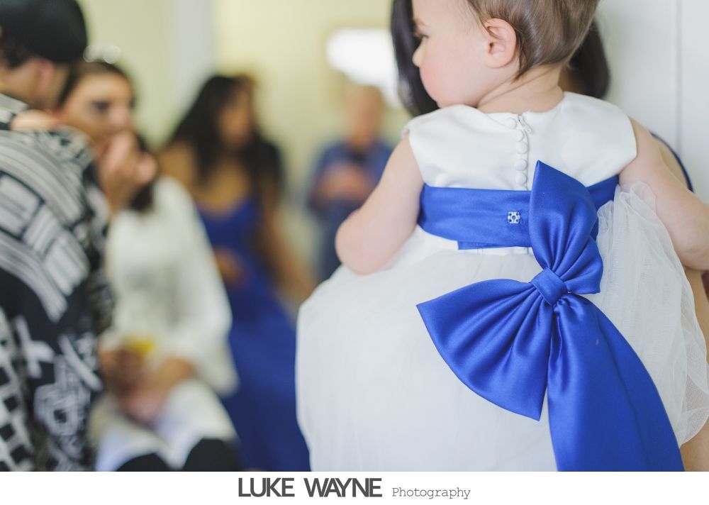 Back view of a baby in a white dress with a large blue bow, indoors.