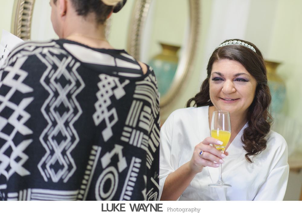 Bride in white robe and tiara, holding mimosa, smiling, with person in black and white patterned top.