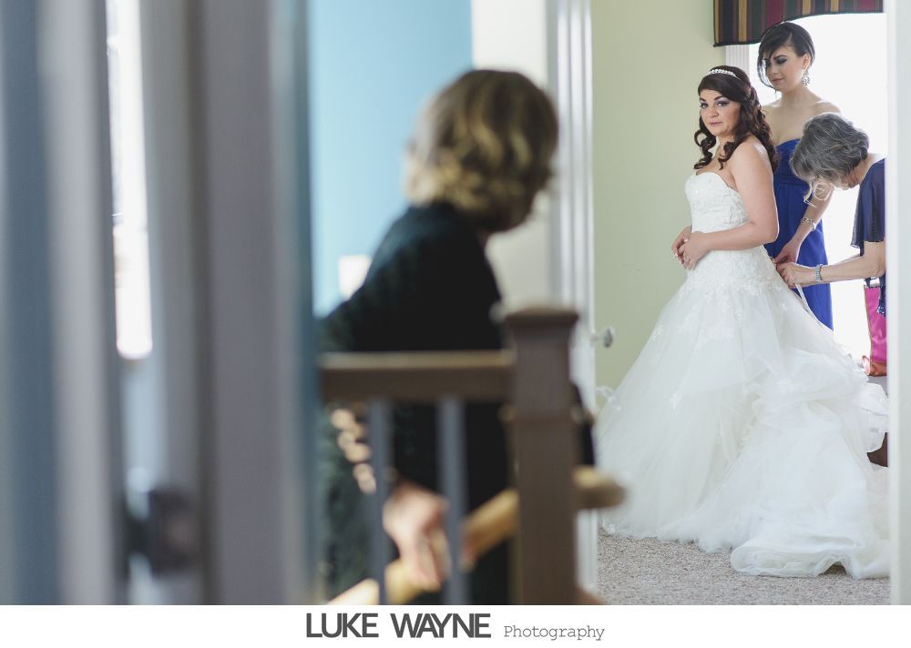 Bride in a white gown being helped by two people. Window and doorway in the background.