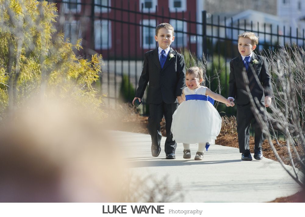 Three children in wedding attire walking on a path. A flower girl in white and two boys in suits.