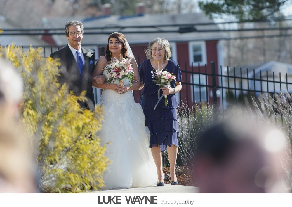 Bride walking down an aisle, arm-in-arm with her parents. Wedding ceremony outdoors.