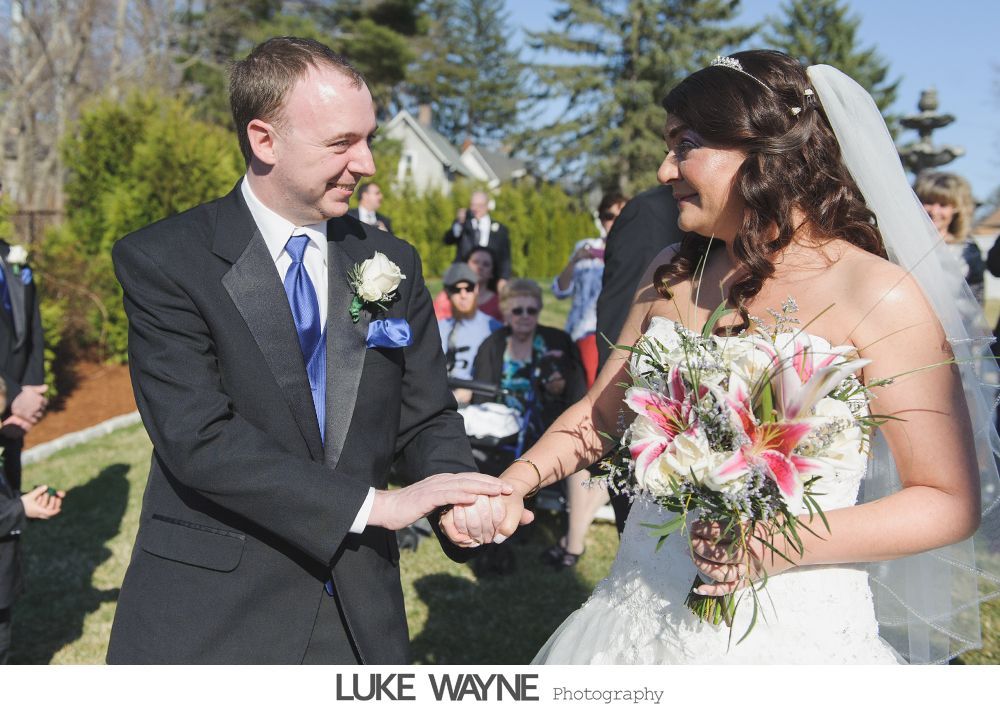 Groom in black tux and bride in white dress holding hands outdoors, smiling at each other.