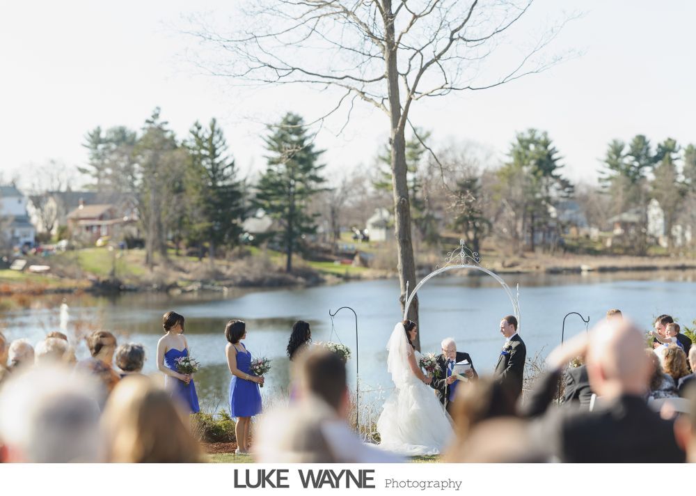 Wedding ceremony by a lake; bride and groom under an arch, bridesmaids in blue, guests seated, sunny day.