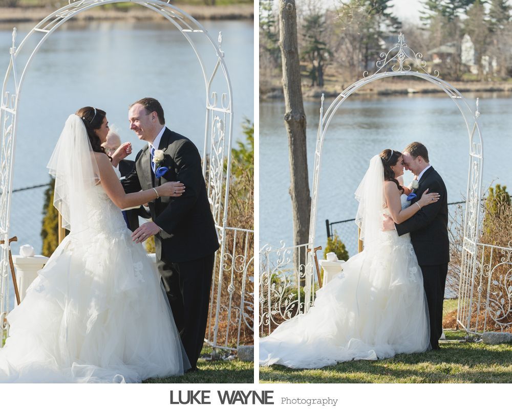 Bride and groom embrace under an archway overlooking a lake.