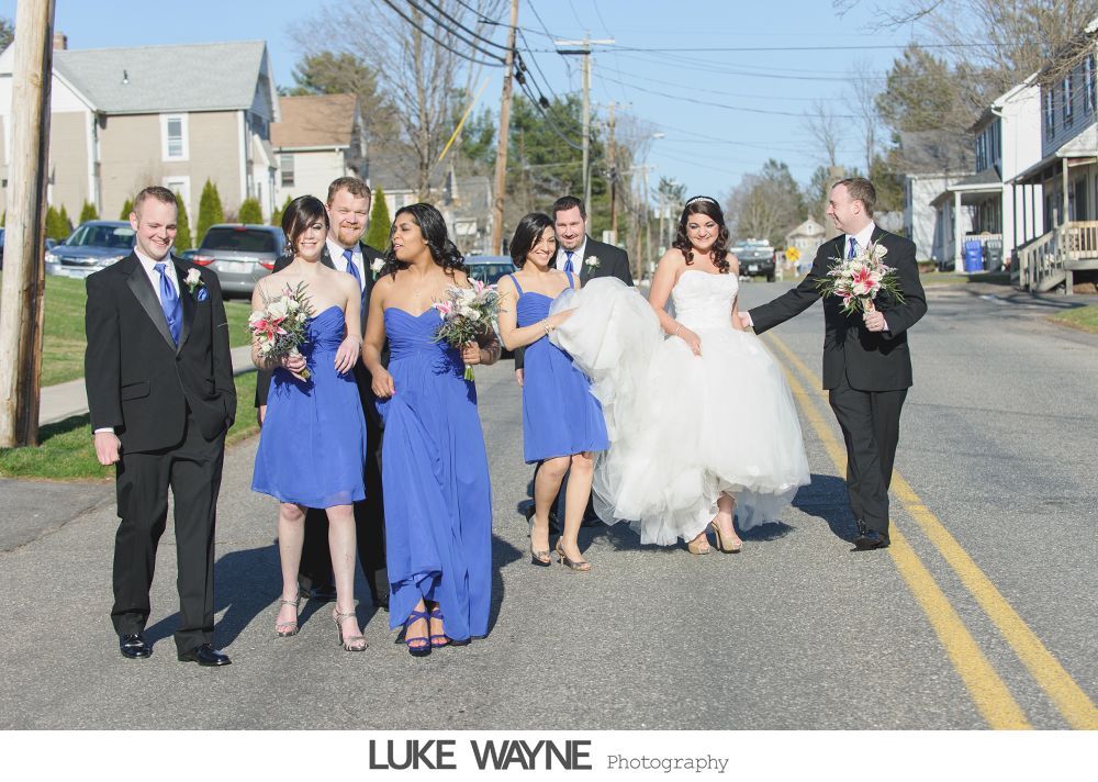 Wedding party walking in street; bride in white gown, bridesmaids in blue dresses, groomsmen in suits.