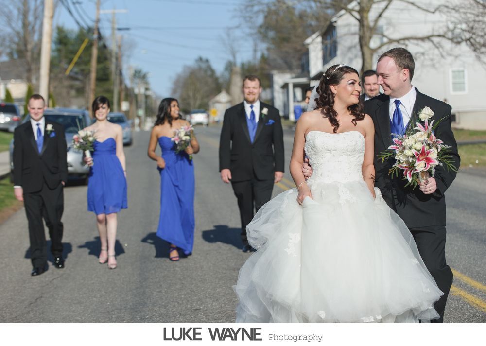 Wedding party walking down a street; bride in white dress, bridesmaids in blue, groomsmen in black suits, all smiling.