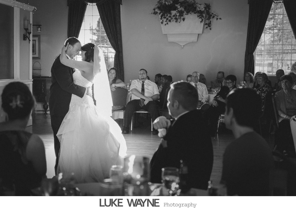 Bride and groom dance at wedding reception, illuminated by window light. Guests watch from tables. Black and white.