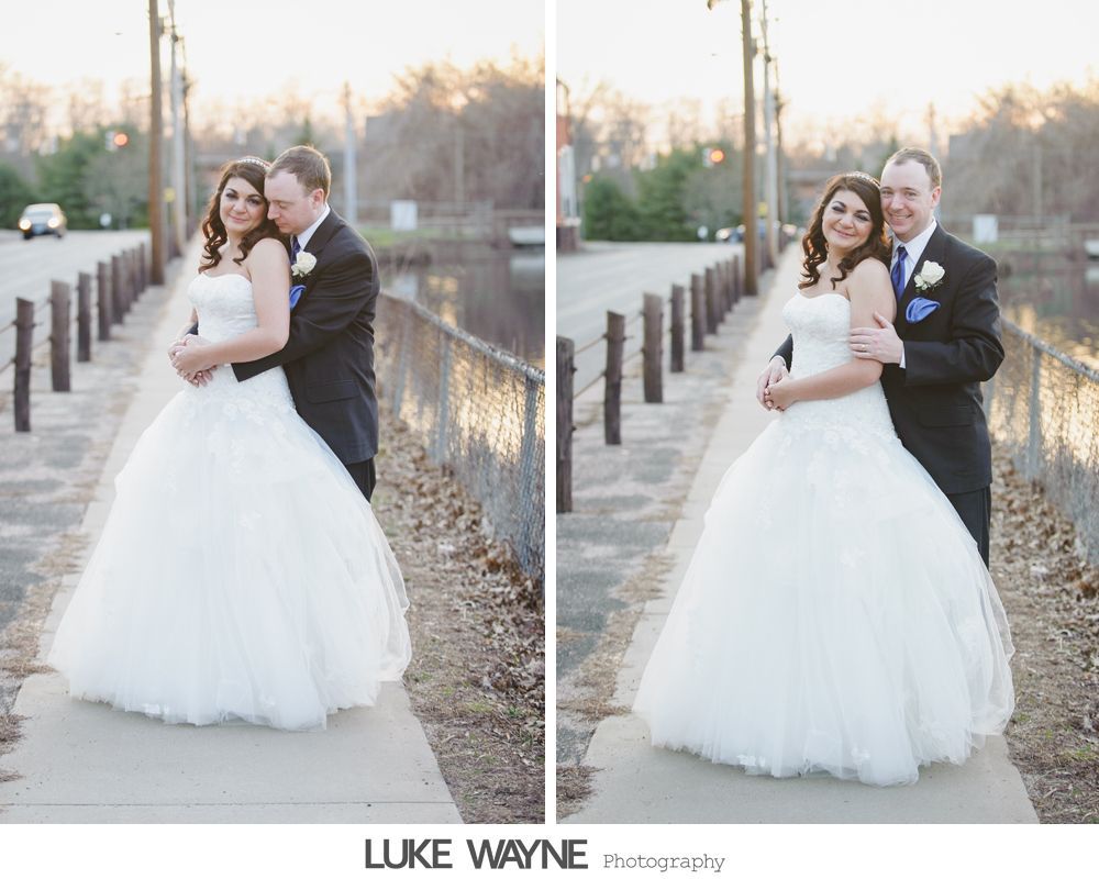 Bride and groom embrace on a pathway next to a waterway. The bride wears a white ball gown; the groom, a dark suit.