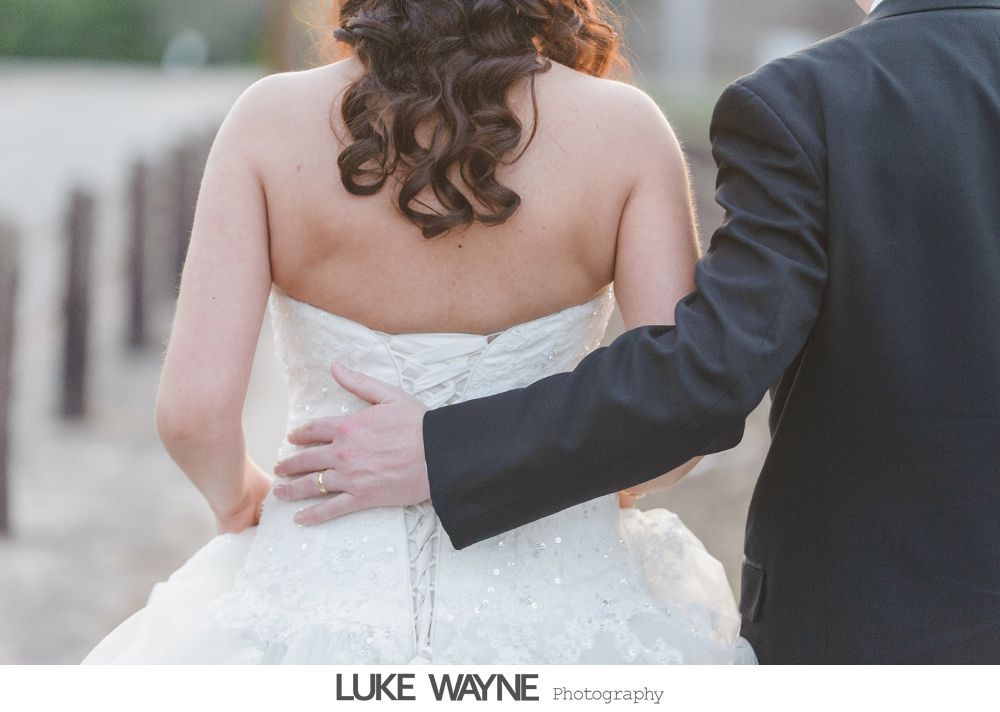 Bride in white strapless dress, groom in black suit jacket, arm around bride's waist, walking outdoors.