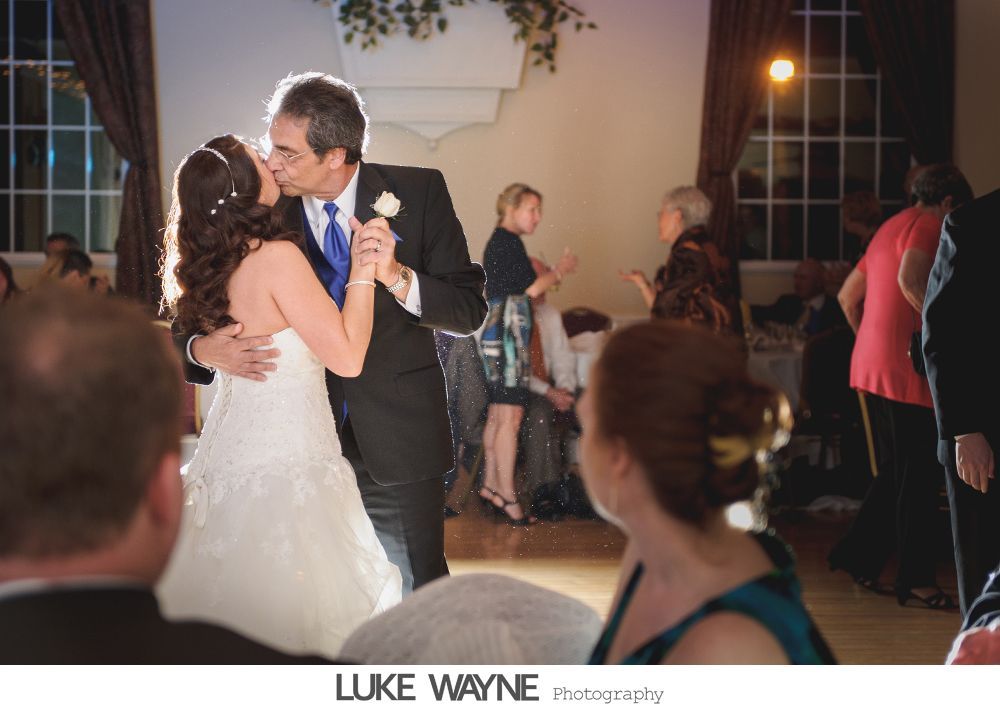 Bride and groom kissing during first dance at wedding reception. Others watch. Warm lighting.