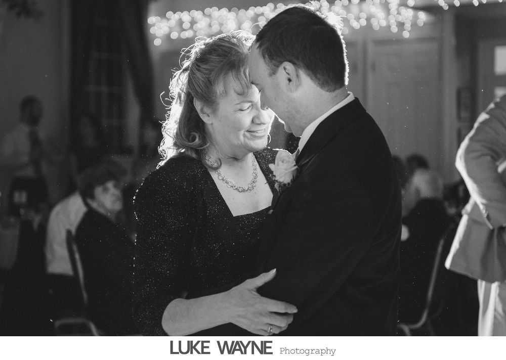Man and woman embrace and dance at wedding reception. Woman smiles with tears, lit by overhead lights.
