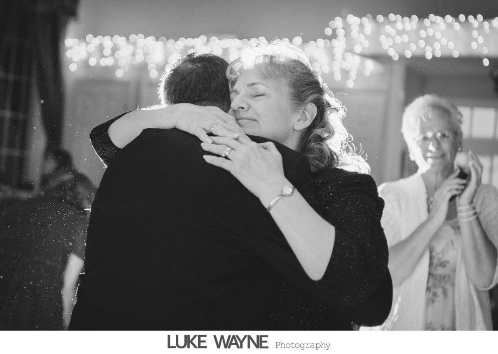 Woman embracing someone while another claps in the background, illuminated by string lights.