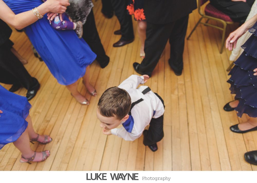 Boy in formal wear dances on wooden floor, surrounded by people in dresses and suits.