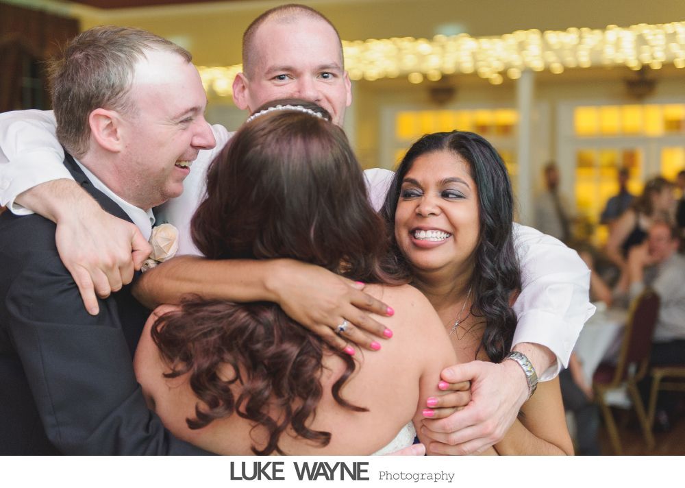 Four people in a group hug at a wedding reception; two men and two women smiling.