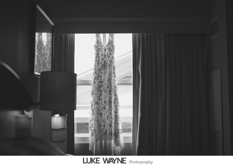 Wedding dress hanging in a bright window, draped against drawn curtains. Black and white.