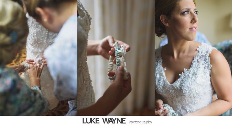 Bride in embellished wedding dress, having it fastened by another. Bride looks away, holding earrings.