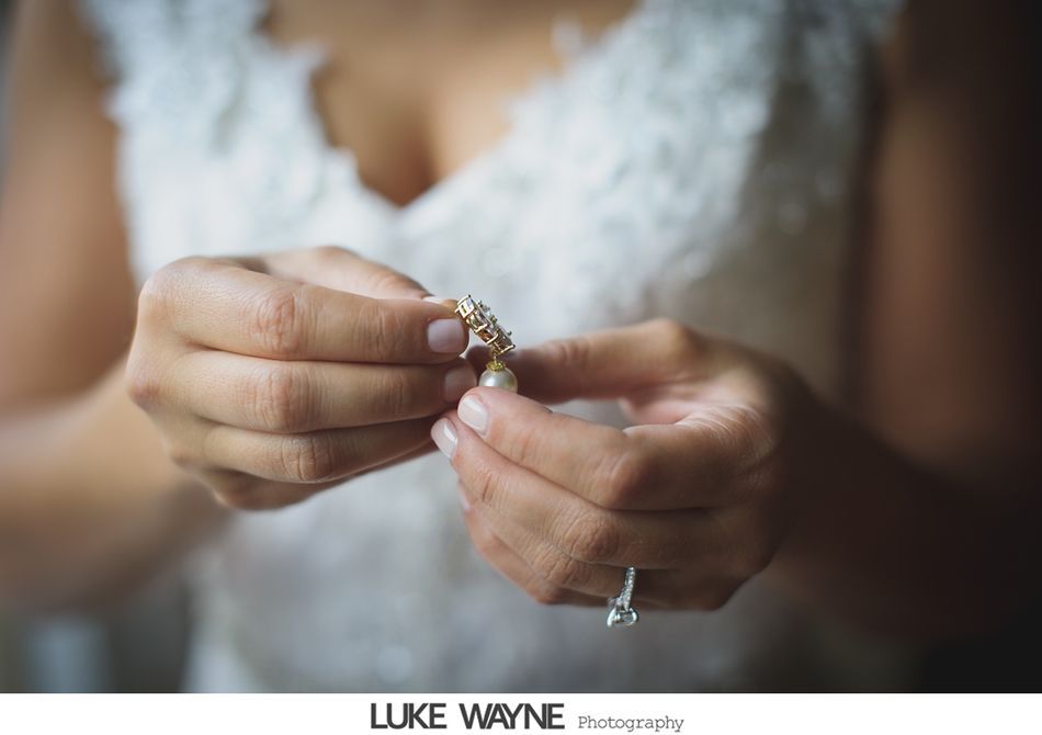 Bride holding a pearl earring, wearing a white lace dress, showing engagement ring.