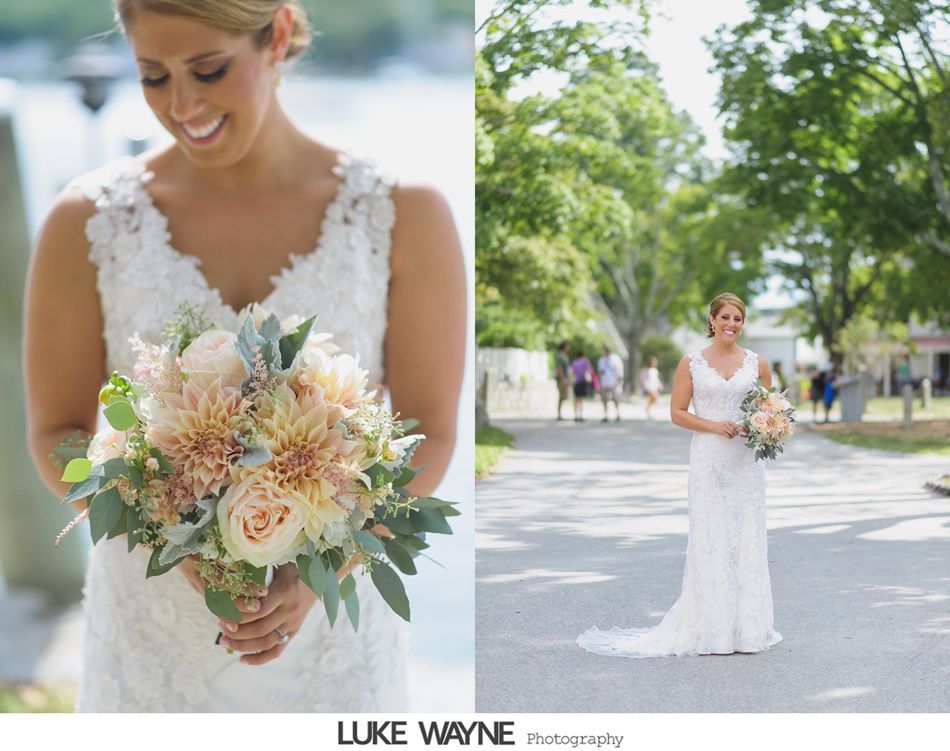 Bride in white lace dress smiles, holding a bouquet of flowers, standing outside on a sunny day.
