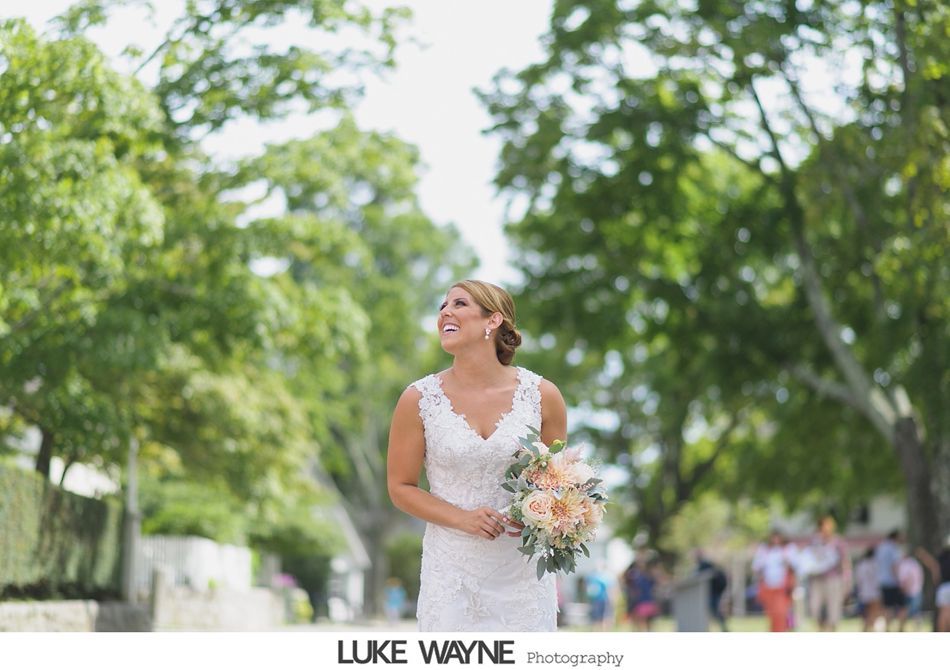 Bride in white lace dress holding flowers, smiles, stands on tree-lined street.