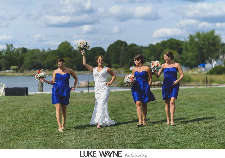 Bride and bridesmaids in blue dresses walking on grass by water; happy expressions.