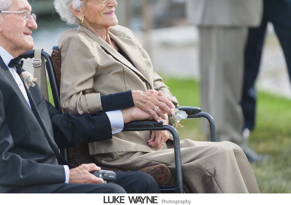 Elderly couple holding hands, one in a wheelchair, at an outdoor event.