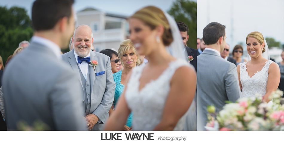 Wedding ceremony with the bride and groom smiling at each other. Guests and a man in a gray suit are visible.