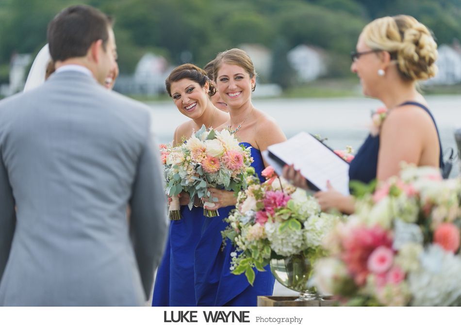 Wedding ceremony, bridesmaids smiling at the groom, officiant reading, ocean background.