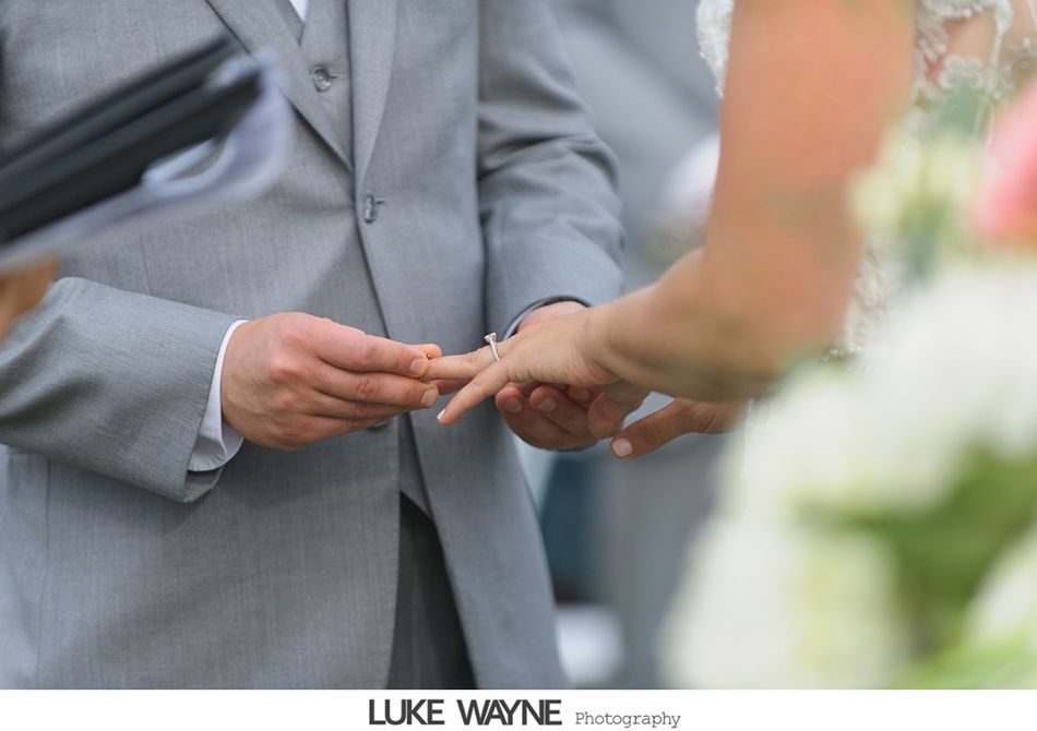 Groom placing a ring on bride's finger during a wedding ceremony. Groom wears a gray suit, bride's arm is visible.