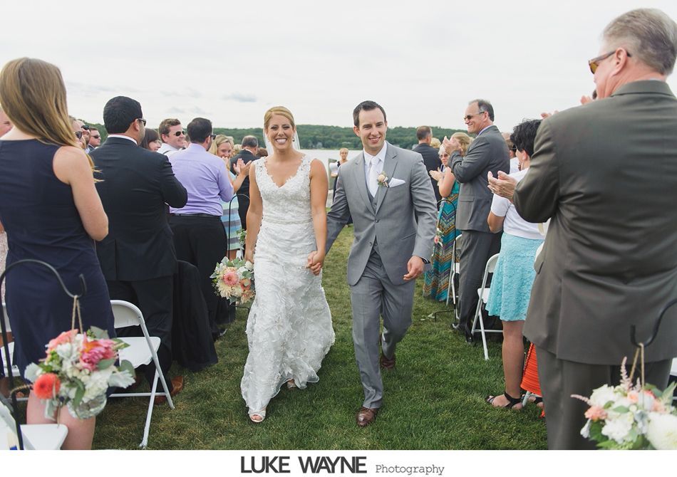 Newlyweds walking down the aisle, smiling, holding hands, surrounded by applauding guests outdoors.