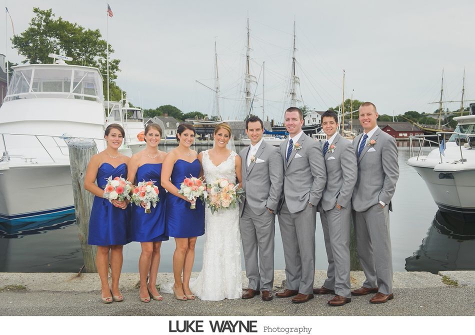 Wedding party by a harbor: bride, bridesmaids in blue, groomsmen in gray suits, boats, and sailboats.