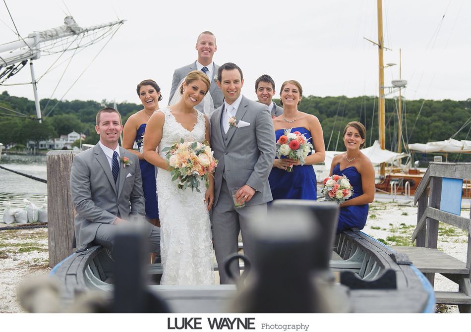 Wedding party poses on a boat in front of a harbor. The bride and groom are in the center, surrounded by bridesmaids in blue and groomsmen in gray.