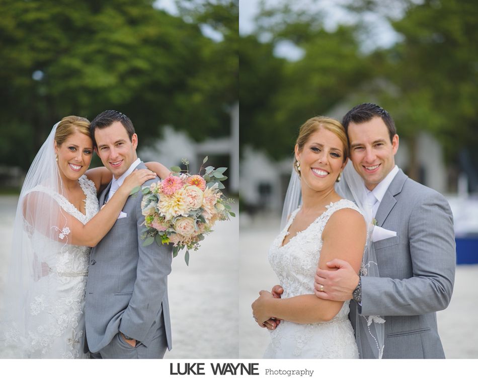 Bride and groom embrace, smiling. She wears a white lace dress and veil; he wears a gray suit. Outdoors.