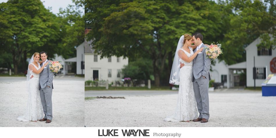 Newlyweds embrace, holding bouquet in front of a white building with trees in the background.