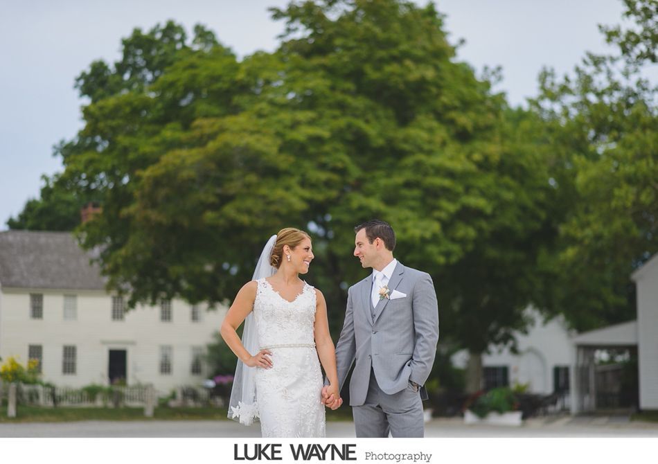 Bride and groom holding hands, smiling at each other. They stand outside near a tree and buildings.
