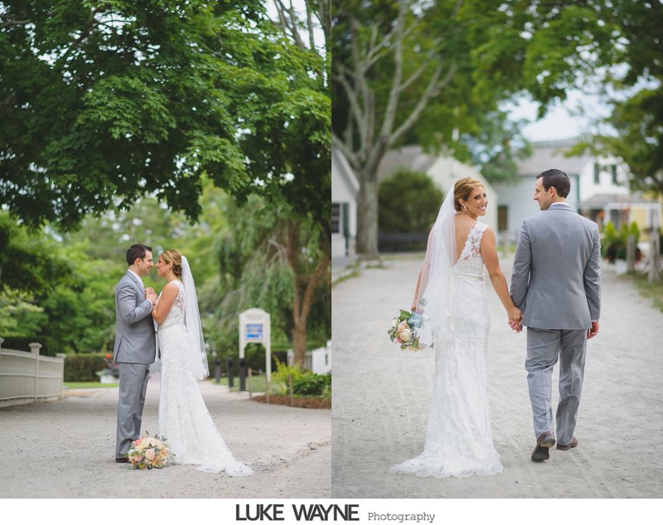Bride and groom in formal wear hold hands and smile while walking on a path lined with trees and buildings.