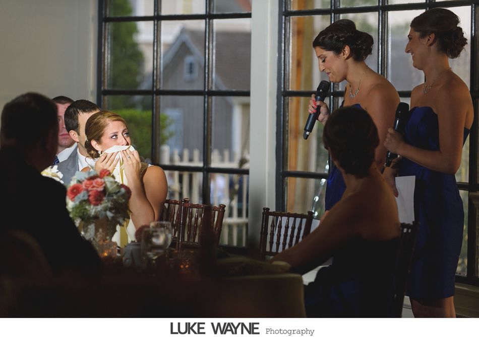 Bride wiping tears during a toast. Two women in blue dresses stand nearby with microphones.