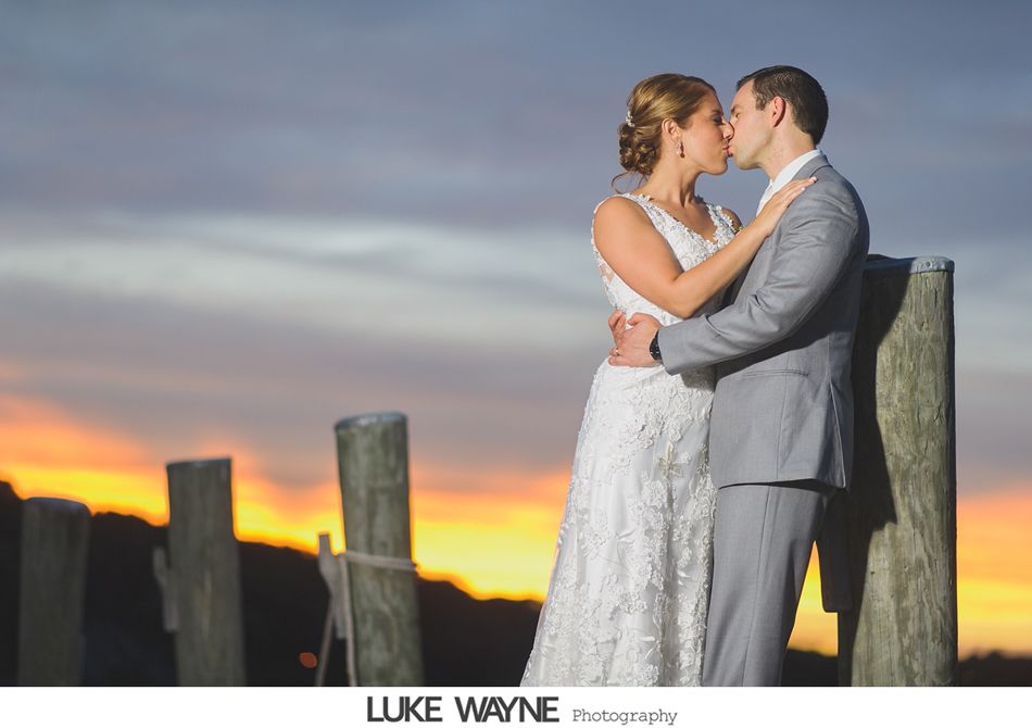 Couple kissing at sunset on a dock; bride in white lace dress, groom in gray suit.