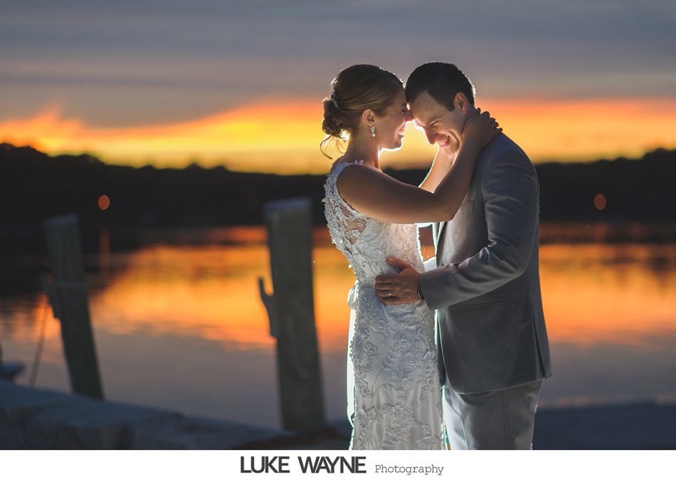 Couple embraces at sunset over water. Bride in white dress, groom in grey suit.