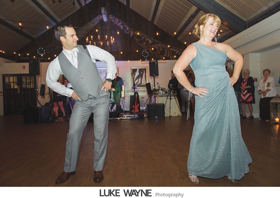 Couple dancing at a reception, posing with hands on hips. Man in grey suit, woman in teal gown. Wooden floor, string lights.