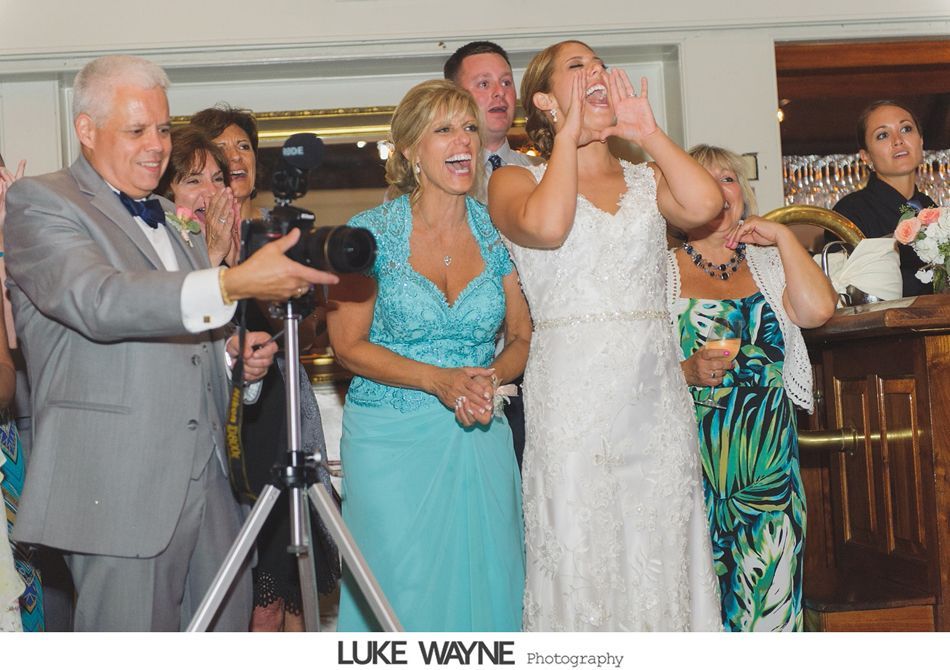 Wedding guests cheer, a bride yells, photographer captures the moment indoors, warm lighting.